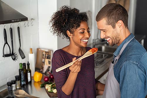 An attractive young couple cooking dinner together. She is having him taste the sauce she's making
